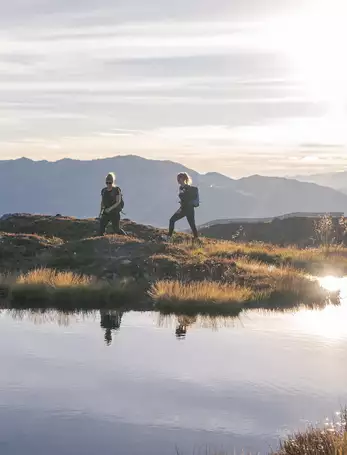 Zwei Frauen wandern bei einem kleinen Bergsee vorbei, bei Sonnenschein