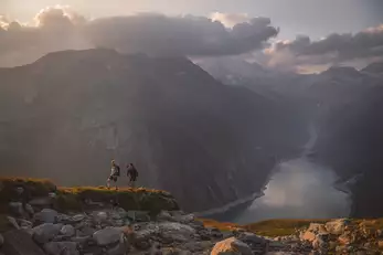 Zwei Wanderer sind auf dem Weg zur Hütte, im Hintergrund sieht man einen Speicherteich