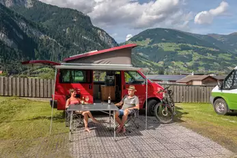 Ein Camper Paar sitzt vor einem roten Camper beim Wein- und Biertrinken