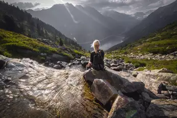 Frau sitzt bei einem Bach und schaut auf das Bergpanorama mit Speichersee